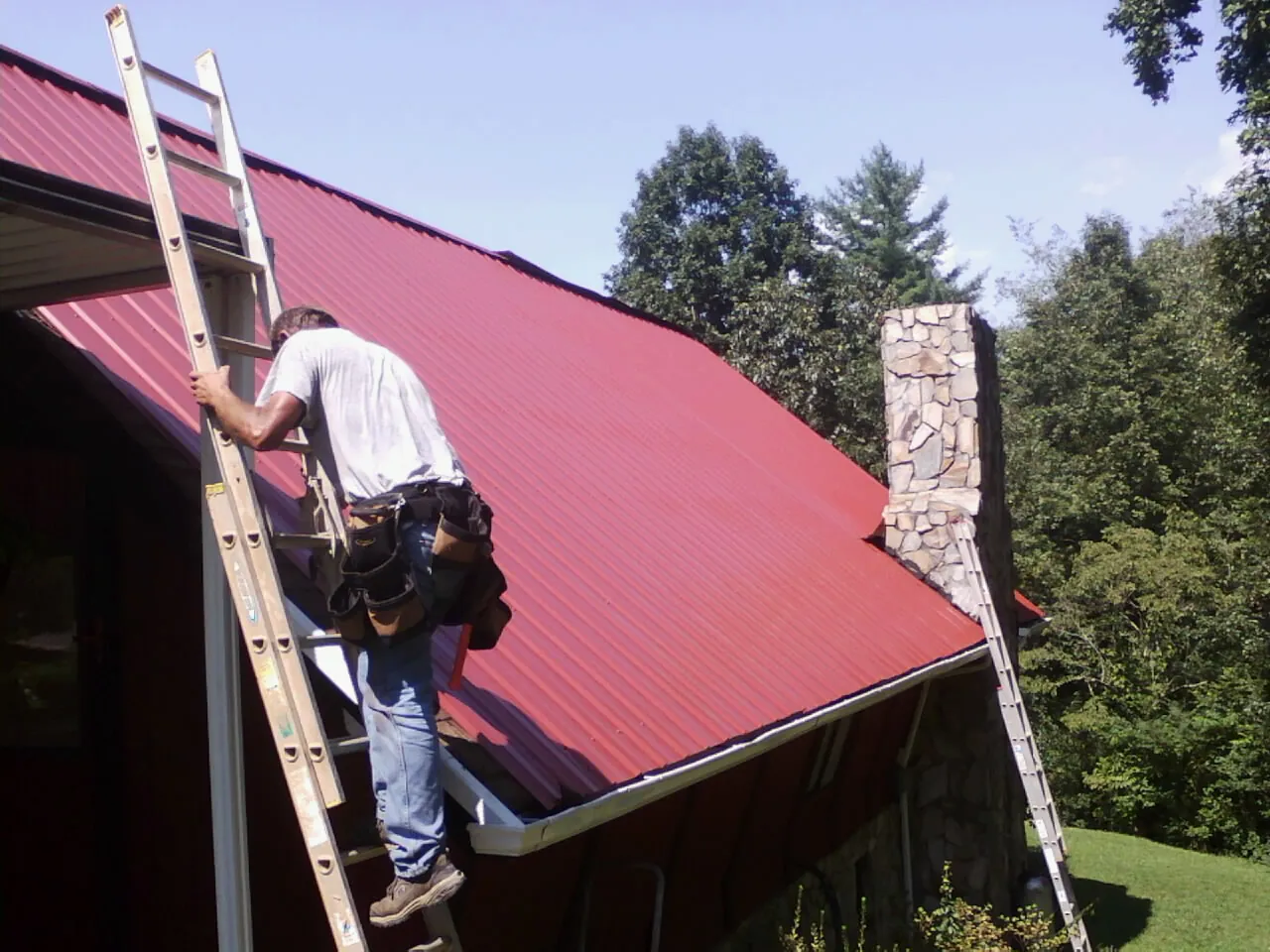 Roofer on a ladder beside a red metal roof and stone chimney — Soffit & Fascia Repair by Roofing Brothers in Rockwood